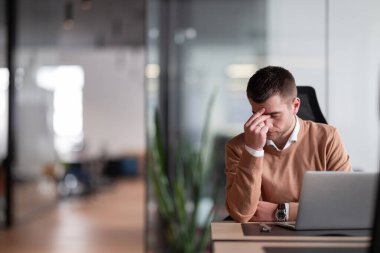 Ofiste dizüstü bilgisayar üzerinde çalışırken baş ağrısı, yorgunluk, yorgunluk ve stres yaşayan bir iş adamı. Belirtileri arasında migren, depresyon, anksiyete, baş dönmesi, beyin sisi ve zihinsel sağlık sorunları yer alıyor.