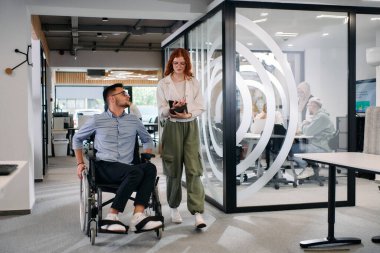 İş arkadaşları, işbirlikçi iş arkadaşları, tekerlekli sandalyedeki bir kişi de dahil olmak üzere, iş yerlerindeki çeşitliliği, takım çalışmasını ve yetkilendirmeyi gösteren modern cam ofis koridorunun yanından geçtiler.