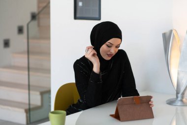 Kendine güveni tam, siyah başörtülü genç bir Müslüman kadın modern bir masada tablet, fincan ve dekoratif lambayla modern bir iç mekanda oturuyor.. 