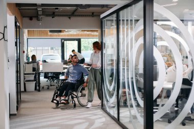 İş arkadaşları, işbirlikçi iş arkadaşları, tekerlekli sandalyedeki bir kişi de dahil olmak üzere, iş yerlerindeki çeşitliliği, takım çalışmasını ve yetkilendirmeyi gösteren modern cam ofis koridorunun yanından geçtiler.