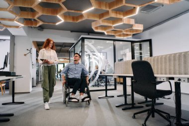 İş arkadaşları, işbirlikçi iş arkadaşları, tekerlekli sandalyedeki bir kişi de dahil olmak üzere, iş yerlerindeki çeşitliliği, takım çalışmasını ve yetkilendirmeyi gösteren modern cam ofis koridorunun yanından geçtiler.