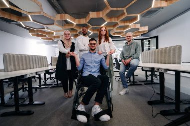 Modern bir ofisteki genç işadamları, tekerlekli sandalyedeki iş arkadaşlarına el sıkışarak, şirket ortamında kapsayıcılık, destek ve birlik gösteriyorlar.