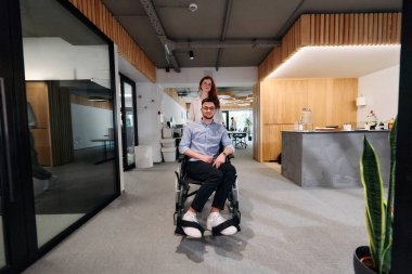 İş arkadaşları, işbirlikçi iş arkadaşları, tekerlekli sandalyedeki bir kişi de dahil olmak üzere, iş yerlerindeki çeşitliliği, takım çalışmasını ve yetkilendirmeyi gösteren modern cam ofis koridorunun yanından geçtiler.