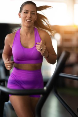 Spor salonunda Koşu bandında Koşan Kadın. Mor spor kıyafeti giyen fit bir kadın spor salonundaki koşu bandında koşuyor. Odaklanmış ama rahat bir ifadesi var. Spor aletleri ve sıcak bir ışığı var.