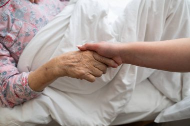 Genç bir kadın, hastane yatağında yaşlı bir kadının ellerini nazikçe tutar. Bakımı, desteği ve merhameti simgeler.