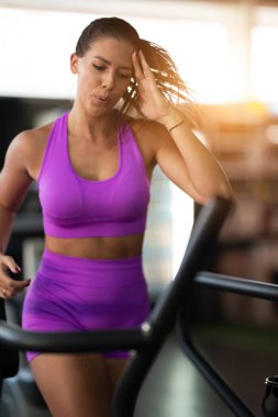 Spor salonunda Koşu bandında Koşan Kadın. Mor spor kıyafeti giyen fit bir kadın spor salonundaki koşu bandında koşuyor. Odaklanmış ama rahat bir ifadesi var. Spor aletleri ve sıcak bir ışığı var.