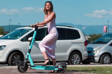 Pembe elbiseli ve güneş gözlüklü zarif bir kadın güneşli şehir sokaklarında elektrikli scooterla geziyor. Mükemmel bir şehir macerasının özgürlüğünü ve tarzını kucaklıyor.