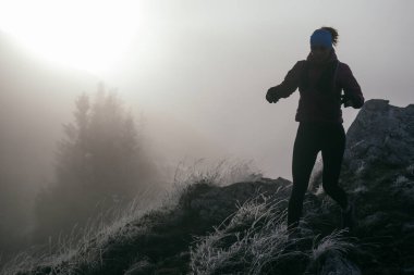 Soğuk ve sisli havada kayalık bir dağ yolunda koşan bir kadın sporcu, sıcak bir ceket, eldiven ve saç bandı giyiyor, dayanıklılık ve kararlılık sergiliyor.. 