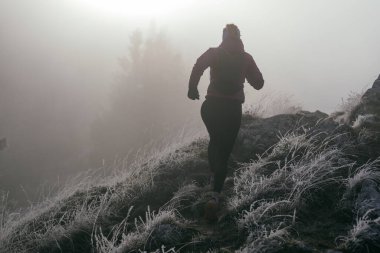 Soğuk ve sisli havada kayalık bir dağ yolunda koşan bir kadın sporcu, sıcak bir ceket, eldiven ve saç bandı giyiyor, dayanıklılık ve kararlılık sergiliyor.. 