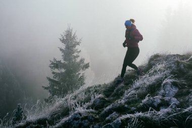 Soğuk ve sisli havada kayalık bir dağ yolunda koşan bir kadın sporcu, sıcak bir ceket, eldiven ve saç bandı giyiyor, dayanıklılık ve kararlılık sergiliyor.. 