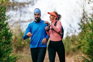 Gülümseyen bir erkek ve kadın, serin bir günde, doğa, fitness ve birbirlerine eşlik ederek, spor giysileri içinde, manzaralı bir orman patikasında birlikte koşarlar..