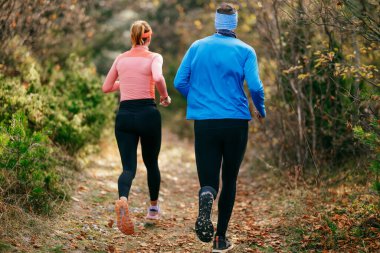 Gülümseyen bir erkek ve kadın, serin bir günde, doğa, fitness ve birbirlerine eşlik ederek, spor giysileri içinde, manzaralı bir orman patikasında birlikte koşarlar..