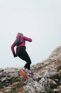 Soğuk ve sisli havada kayalık bir dağ yolunda koşan bir kadın sporcu, sıcak bir ceket, eldiven ve saç bandı giyiyor, dayanıklılık ve kararlılık sergiliyor.. 