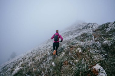 Soğuk ve sisli havada kayalık bir dağ yolunda koşan bir kadın sporcu, sıcak bir ceket, eldiven ve saç bandı giyiyor, dayanıklılık ve kararlılık sergiliyor.. 