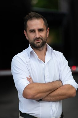 Beyaz gömlekli başarılı bir iş adamı, kollarını kavuşturmuş, açık havada poz veriyor, yüzünde kendinden emin bir ifade var..