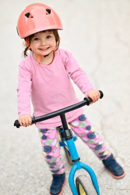 Şirin bir kostüm giymiş ve kask takan küçük bir kız heyecanlı bir şekilde ilk kez bisiklet sürmeyi öğreniyor, neşe ve kararlılıkla dolu.