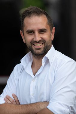 Beyaz gömlekli başarılı bir iş adamı, kollarını kavuşturmuş, açık havada poz veriyor, yüzünde kendinden emin bir ifade var..
