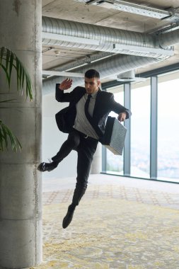 Modern bir ofise kendinden geçmiş bir şekilde atlayan bir işadamı, elinde bir evrak çantasıyla, başarı ve zafer anını kutluyor.