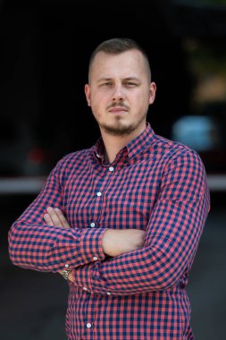 Tişörtlü başarılı genç bir iş adamı, kollarını kavuşturmuş, açık havada poz veriyor, yüzünde kendinden emin bir ifade var.