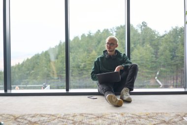 Modern bir ofisin zemininde oturan sarışın bir adam, kucağında dizüstü bilgisayarla pencereye yaslanmış, rahat ve odaklanmış bir tavır sergilerken tamamen işine odaklanmış.