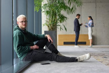 Modern bir ofiste geniş bir pencerenin yanında oturan sıradan bir profesyonel bilgisayarla çalışırken gülümseyen meslektaşları bulanık arka planda etkileşime giren biri.