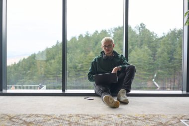 Modern bir ofisin zemininde oturan sarışın bir adam, kucağında dizüstü bilgisayarla pencereye yaslanmış, rahat ve odaklanmış bir tavır sergilerken tamamen işine odaklanmış.