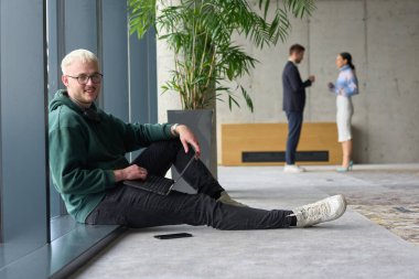 Modern bir ofiste geniş bir pencerenin yanında oturan sıradan bir profesyonel bilgisayarla çalışırken gülümseyen meslektaşları bulanık arka planda etkileşime giren biri.