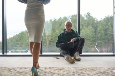 Sarışın bir adam modern bir ofisin zemininde rastgele oturuyor, dizüstü bilgisayarını kullanırken pencereye yaslanıyor, yanında çekici bayan arkadaşı duruyor..