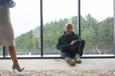 Sarışın bir adam modern bir ofisin zemininde rastgele oturuyor, dizüstü bilgisayarını kullanırken pencereye yaslanıyor, yanında çekici bayan arkadaşı duruyor..