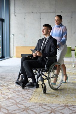Profesyonel bir ofis ortamında, bir iş kadını tekerlekli sandalyede oturan ve tablet kullanan yönetmenine yardım ederken, meslektaşları arka planda işbirliği yapıyor.