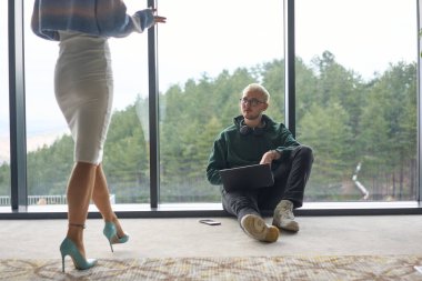 Sarışın bir adam modern bir ofisin zemininde rastgele oturuyor, dizüstü bilgisayarını kullanırken pencereye yaslanıyor, yanında çekici bayan arkadaşı duruyor..