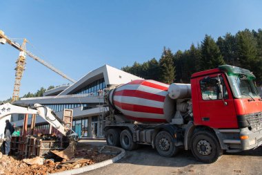 İnşaat halindeki bir iş binasının dış tarafı kamyonlar, vinçler ve çeşitli inşaat malzemeleriyle çevrili. Geniş bir endüstriyel çalışma sahasının hareketli bir sahnesini oluşturuyor.. 