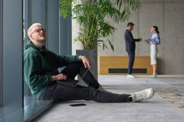 Modern bir ofiste geniş bir pencerenin yanında oturan sıradan bir profesyonel bilgisayarla çalışırken gülümseyen meslektaşları bulanık arka planda etkileşime giren biri.