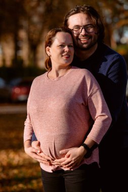 Bir adam parkta hamile karısını sevgiyle kucaklıyor. Huzurlu bir sıcaklık ve şefkat anı yaşıyor..