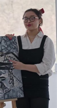 Asyalı kadın sanatçının evde sergilediği dikey video. Önlük takan nüfuzlu, tuvaldeki siyah beyaz bir tabloyu fırçayla işaret ediyor. Resim okulu için mükemmel.