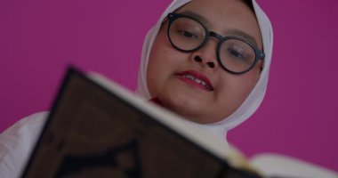 Beyaz başörtüsü takmış, Kuran 'ı Arapça yazan Müslüman bir kadının yakın çekimi. İnancı ve bağlılığı yansıtan kutsal metne derinden odaklanmış durumda. Pembe bir arka planda izole edilmiş..