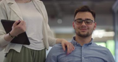 Genç iş arkadaşları, işbirlikçi iş arkadaşları, aralarında tekerlekli sandalyedeki bir kişinin de bulunduğu, modern cam ofis koridorunun yanından geçerek, çeşitliliği, takım çalışmasını ve güçlendirmeyi resmediyorlar.