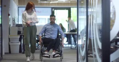 Genç iş arkadaşları, işbirlikçi iş arkadaşları, aralarında tekerlekli sandalyedeki bir kişinin de bulunduğu, modern cam ofis koridorunun yanından geçerek, çeşitliliği, takım çalışmasını ve güçlendirmeyi resmediyorlar.