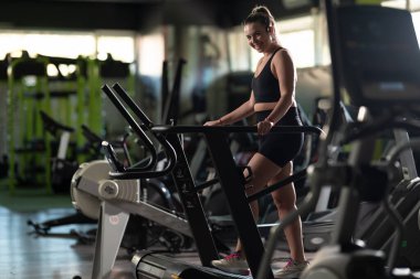 Spor salonunda Koşu bandında Koşan Kadın. Mor spor kıyafeti giyen fit bir kadın spor salonundaki koşu bandında koşuyor. Odaklanmış ama rahat bir ifadesi var. Spor aletleri ve sıcak bir ışığı var.