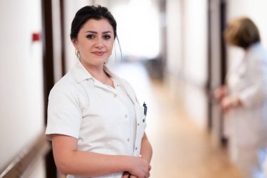 Kendini işine adamış bir hemşire iyi aydınlatılmış bir hastane koridorunda hızla yürüyor. Sağlık hizmetlerindeki profesyonelliği ve aciliyeti yansıtıyor.