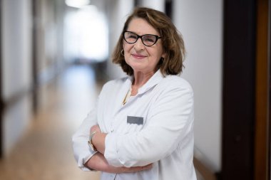 Tecrübeli ve kendinden emin yaşlı bir kadın doktor, gözlük takıyor ve hastane koridorunda duruyor, bilgeliği ve hasta bakımına bağlılığı temsil ediyor.