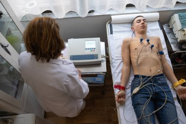 Kadın bir doktor, hastane odasında genç bir adama EKG çeker. Rutin bir kontrol sırasında kalp sağlığını dikkatle izler.