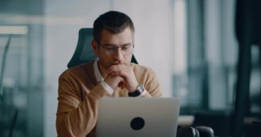 Akıllı görünümlü odaklanmış iş adamı bir dizüstü bilgisayarda çalışıyor ve düşünüyor. Milenyum dönemi aile bütçesini yönetmek, finansal evrak işlerini halletmek ve modern bir açık uzay ofisindeki dizüstü bilgisayardaki harcamaları takip etmek.