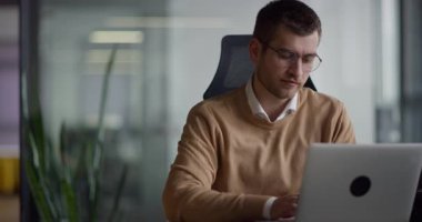 Akıllı görünümlü odaklanmış iş adamı bir dizüstü bilgisayarda çalışıyor ve düşünüyor. Milenyum dönemi aile bütçesini yönetmek, finansal evrak işlerini halletmek ve modern bir açık uzay ofisindeki dizüstü bilgisayardaki harcamaları takip etmek.