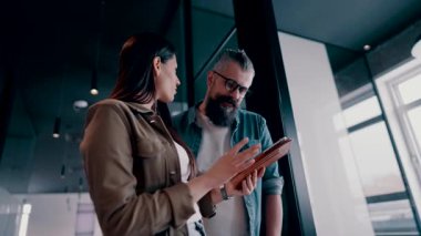 Üst düzey bir hipster akıl hocası ve Avrupalı bir lider, modern bir çalışma alanında tablet üzerinde çalışırken, arka planda yoğun meslektaşlar çalışırken güvenli bir şekilde işbirliği yaparak bir projeyi tartışıyorlar.