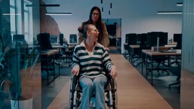 Modern bir ofiste neşeli bir an. Tekerlekli sandalyedeki bir kadın meslektaşı tarafından itilir, ikisi de gülümser. Girişimcilik alanında özel işyeri, takım çalışması, çeşitlilik ve erişilebilirlik
