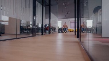 Tekerlekli sandalyede engelli bir adamın olduğu camdan duvarları olan modern bir ofis koridoru. Uzay, parlak, açık dizaynlı ve kapsayıcı bir iş yeri sergiliyor.