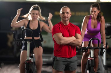 Kararlı bir fitness eğitmeni, iki bayan müşterisinin önünde, arka plandaki antrenmanlarında onlara rehberlik eder.