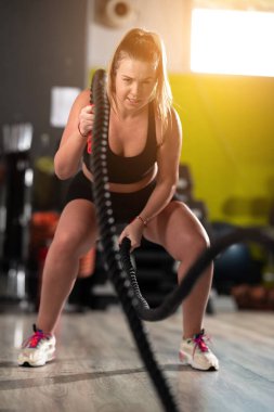 Formda ve atletik bir kadın spor salonunda halatla koşma egzersizleri yapar. Gücünü, dayanıklılığını ve kendini spora adamışlığını gösterir.. 