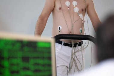 Orta yaşlı bir adam, klinik bir ortamda kalp izleme için EKG elektrotlarına bağlı bir koşu bandında tıbbi stres testinden geçiyor.. 